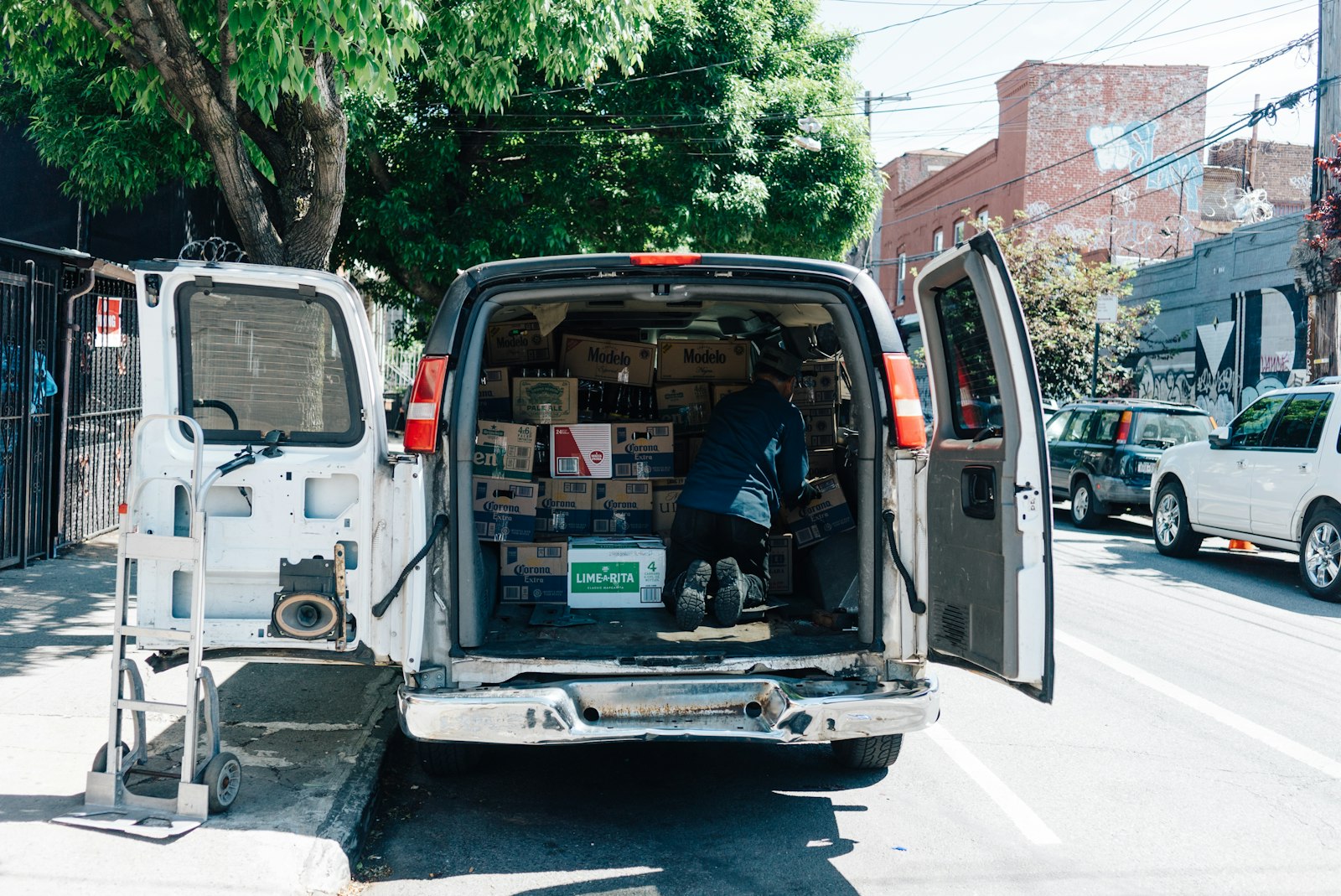 A white cargo van parked kerbside with both rear doors open and a worker stacking boxes inside