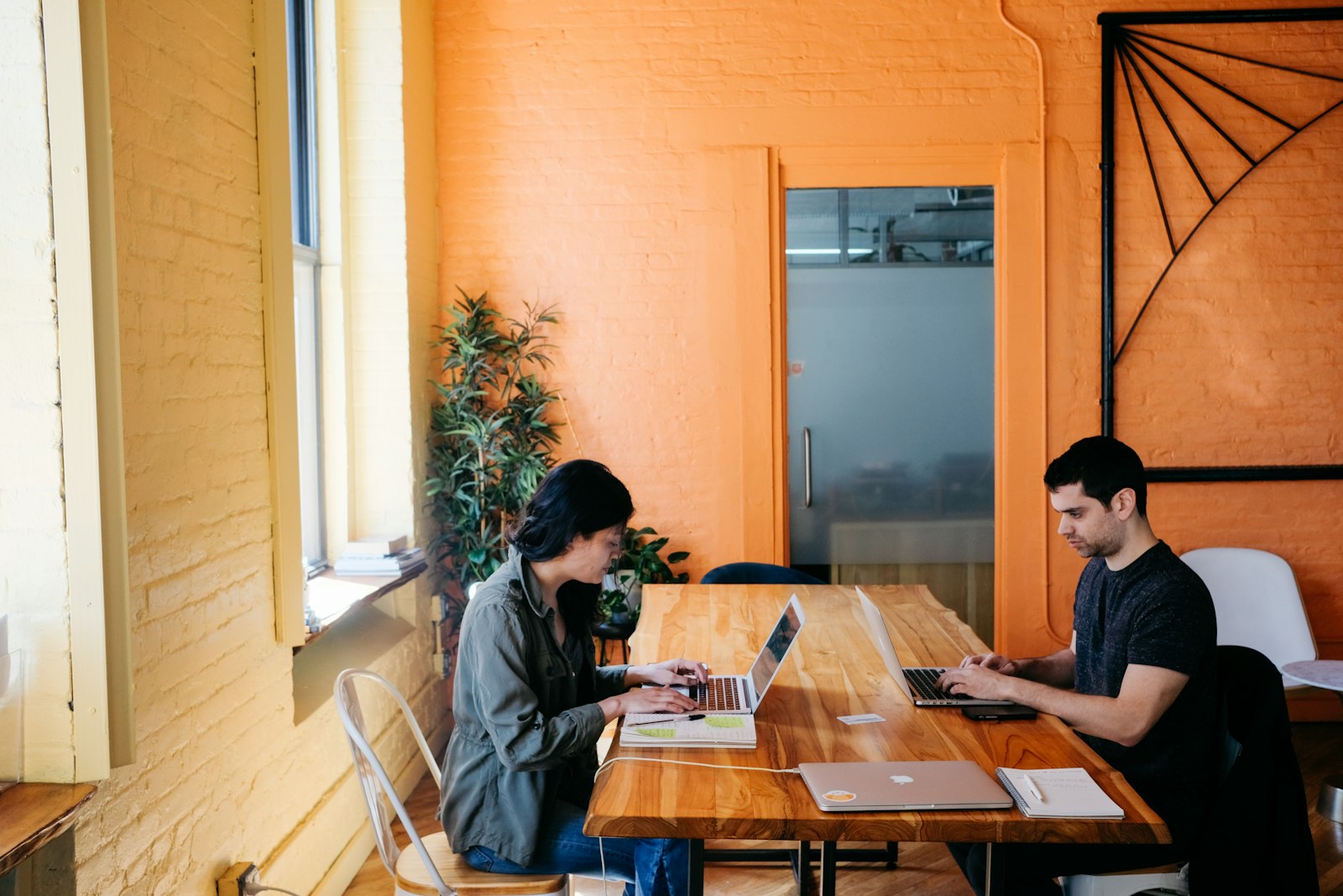 Two colleagues on laptops at a timber table by an orange wall
