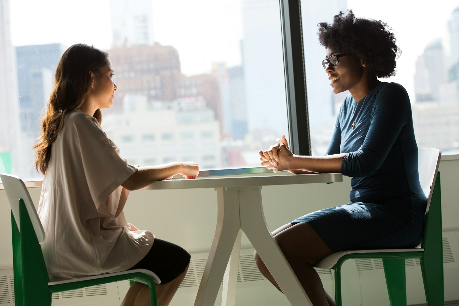 Two women in conversation across a small table by a big window