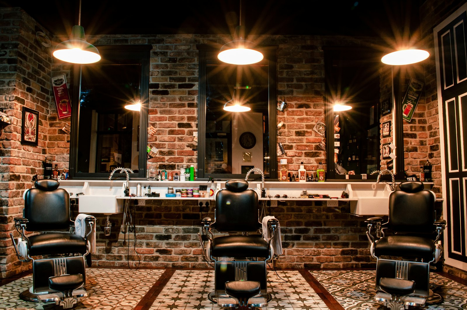 Three black leather barber chairs facing mirrors on an exposed brick wall under pendant lamps