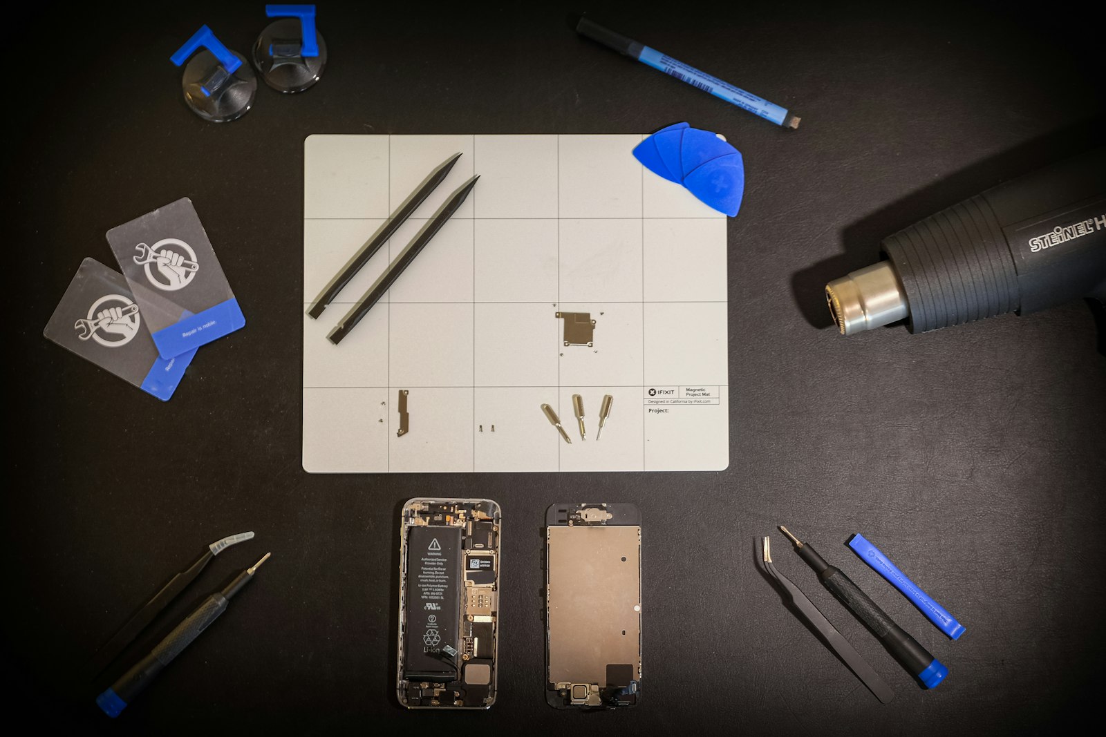A repair kit laid out on black: tools, suction cups, a heat gun and an opened phone
