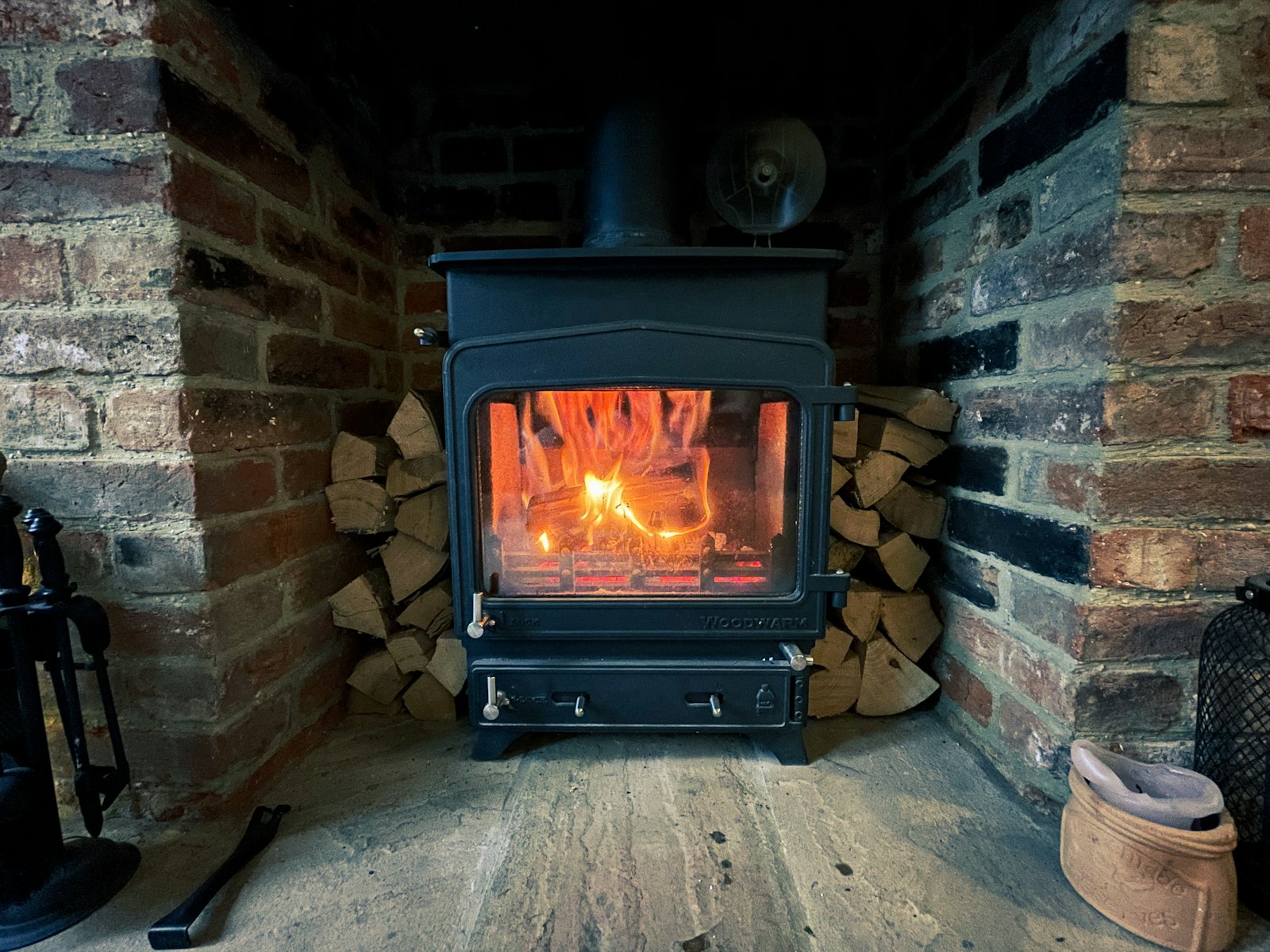 A lit wood burner in a brick fireplace with logs stacked beside it