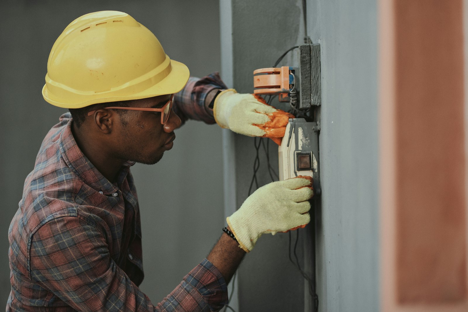 An electrician in a plaid shirt and yellow hard hat wiring a wall socket