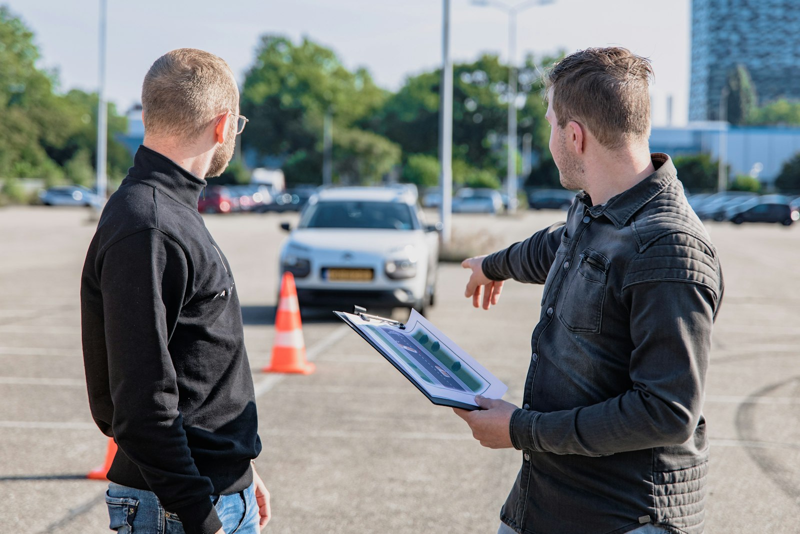 A driving instructor holding a clipboard and pointing towards a car on a practice area