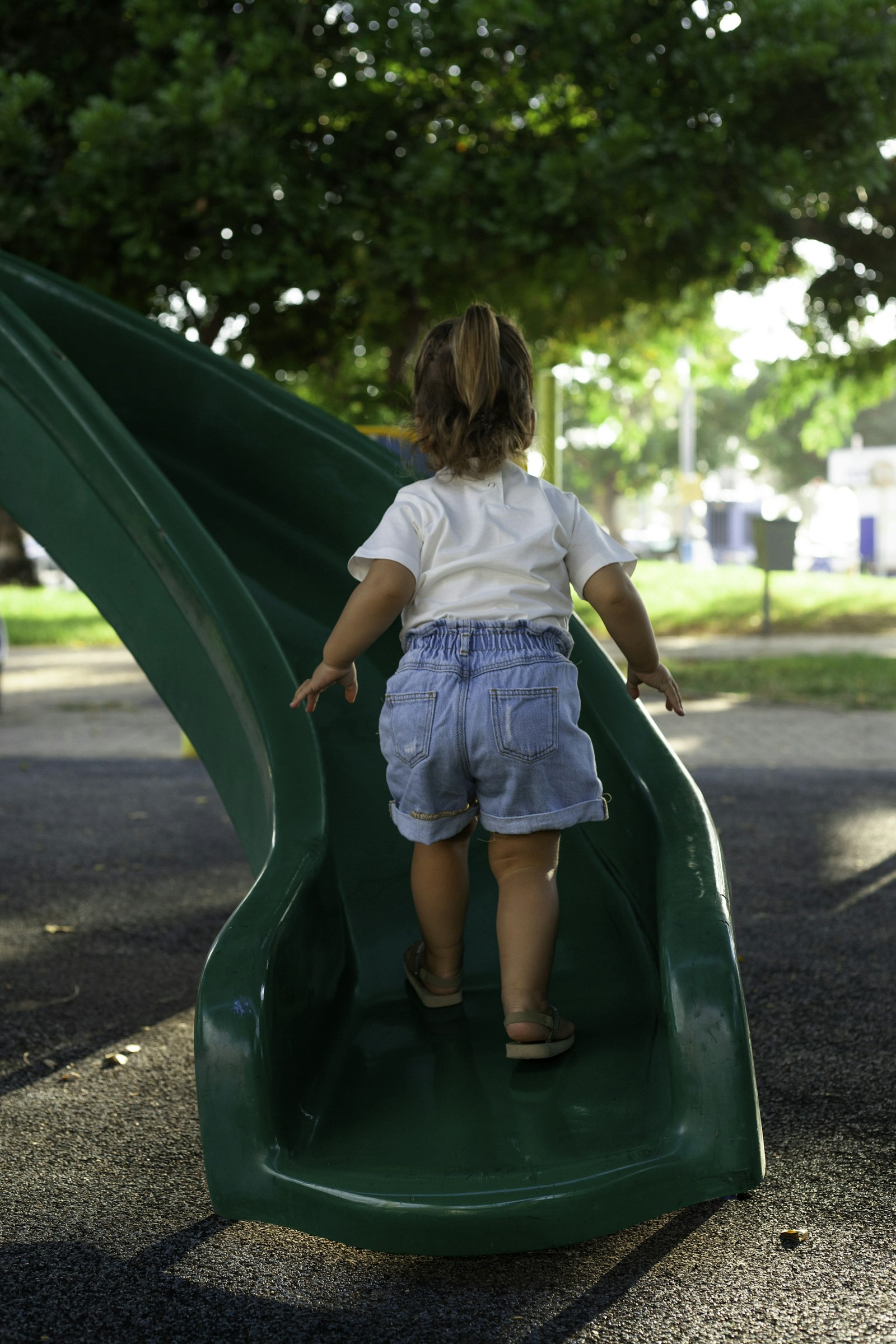 A little girl going down a green slide under trees