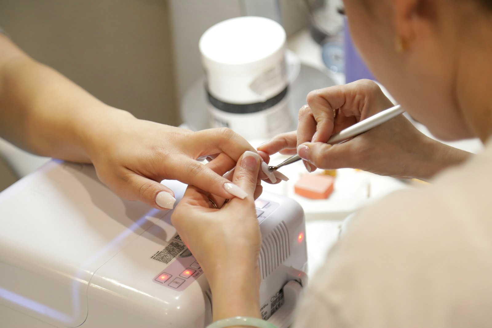 A technician filing a client's nails at a white nail-bar station by the window