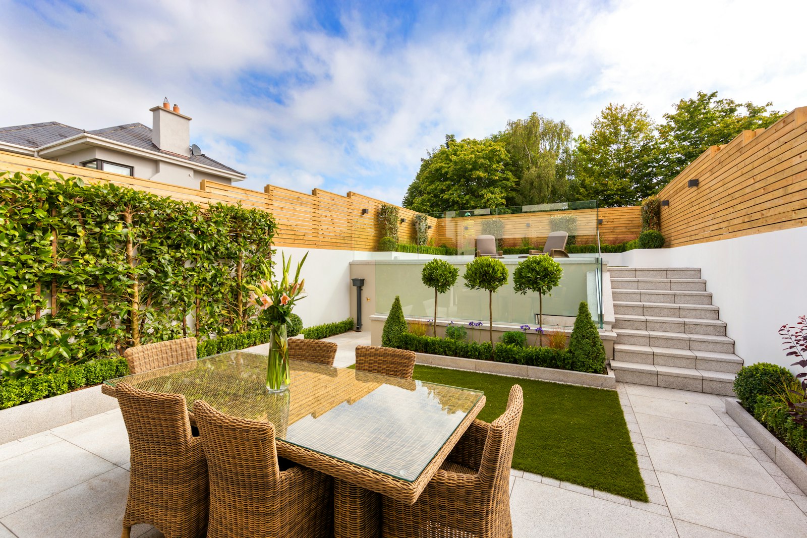 A paved courtyard with a dining table, pleached trees and a strip of lawn