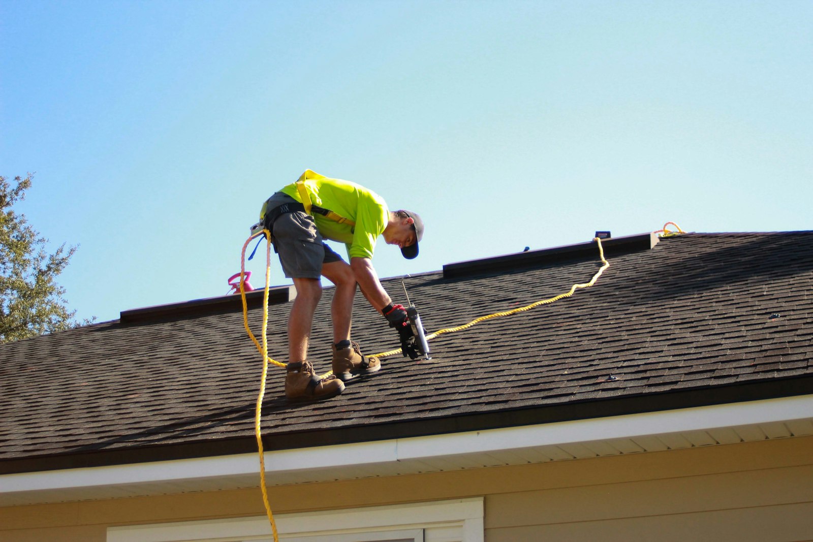 A roofer in a hi-vis shirt and harness drilling shingles on a dark roof