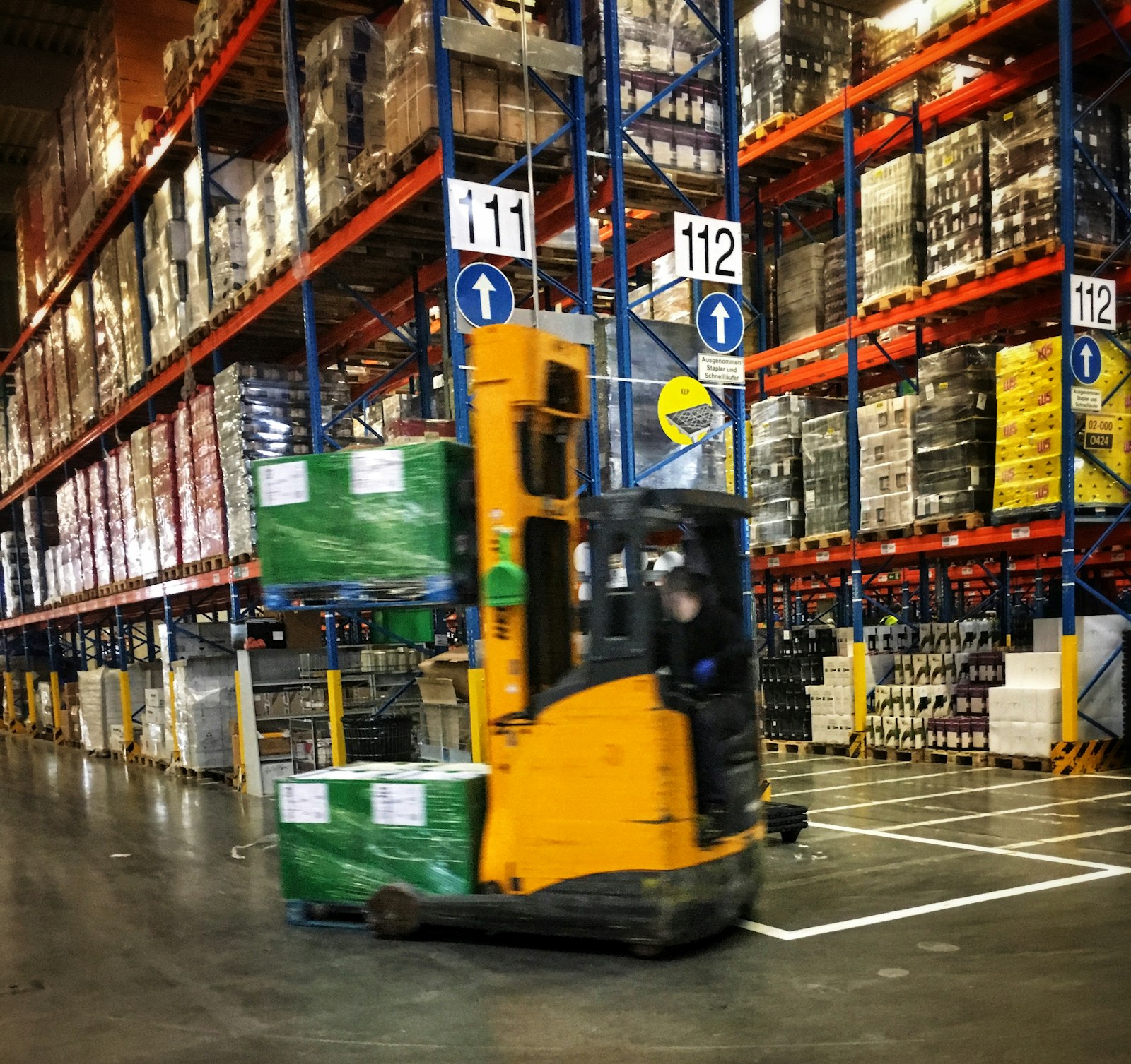 A driver on a yellow reach-truck carrying two shrink-wrapped pallets down a numbered racking aisle