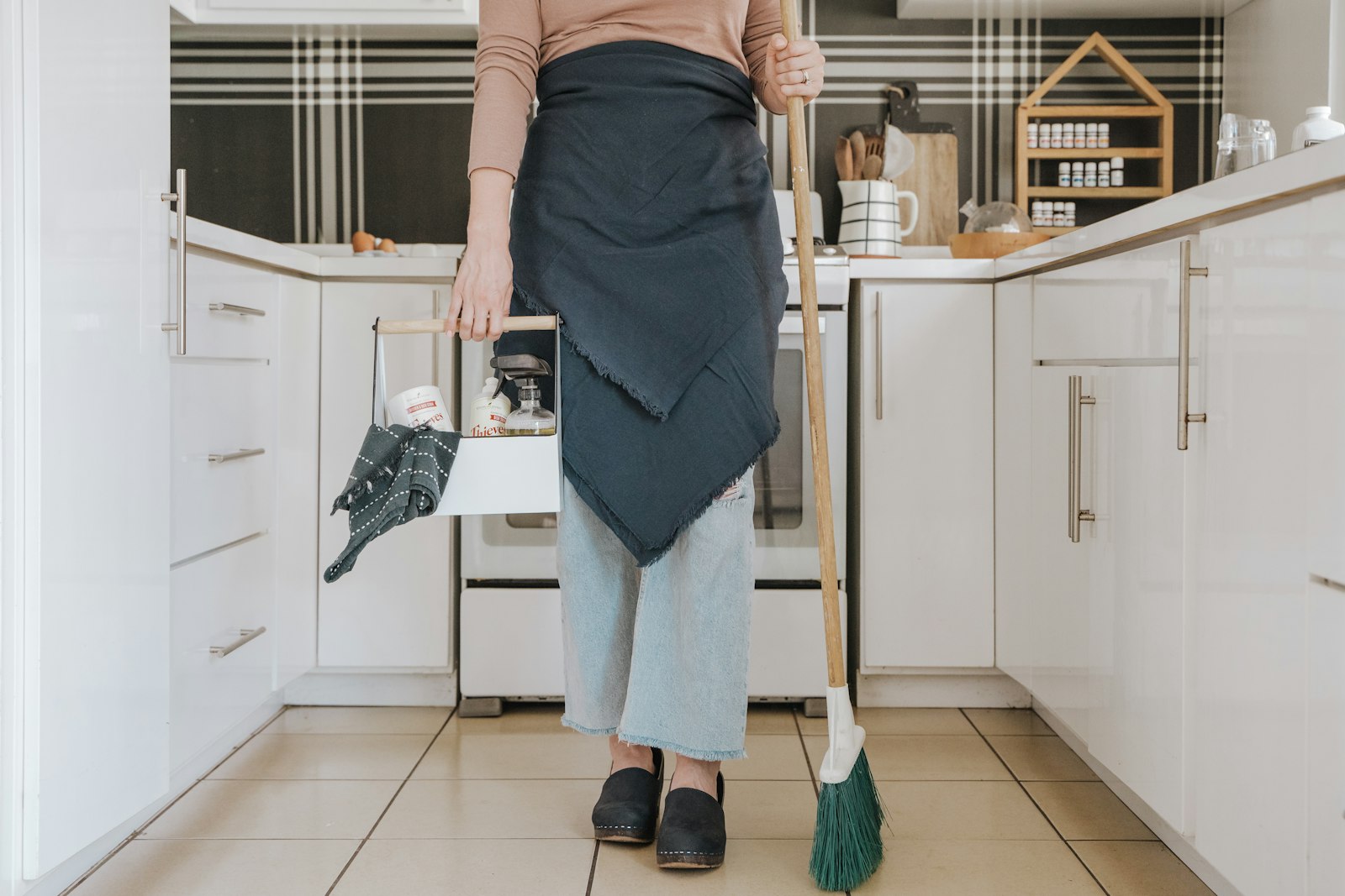 A cleaner in an apron holding a broom in a white kitchen