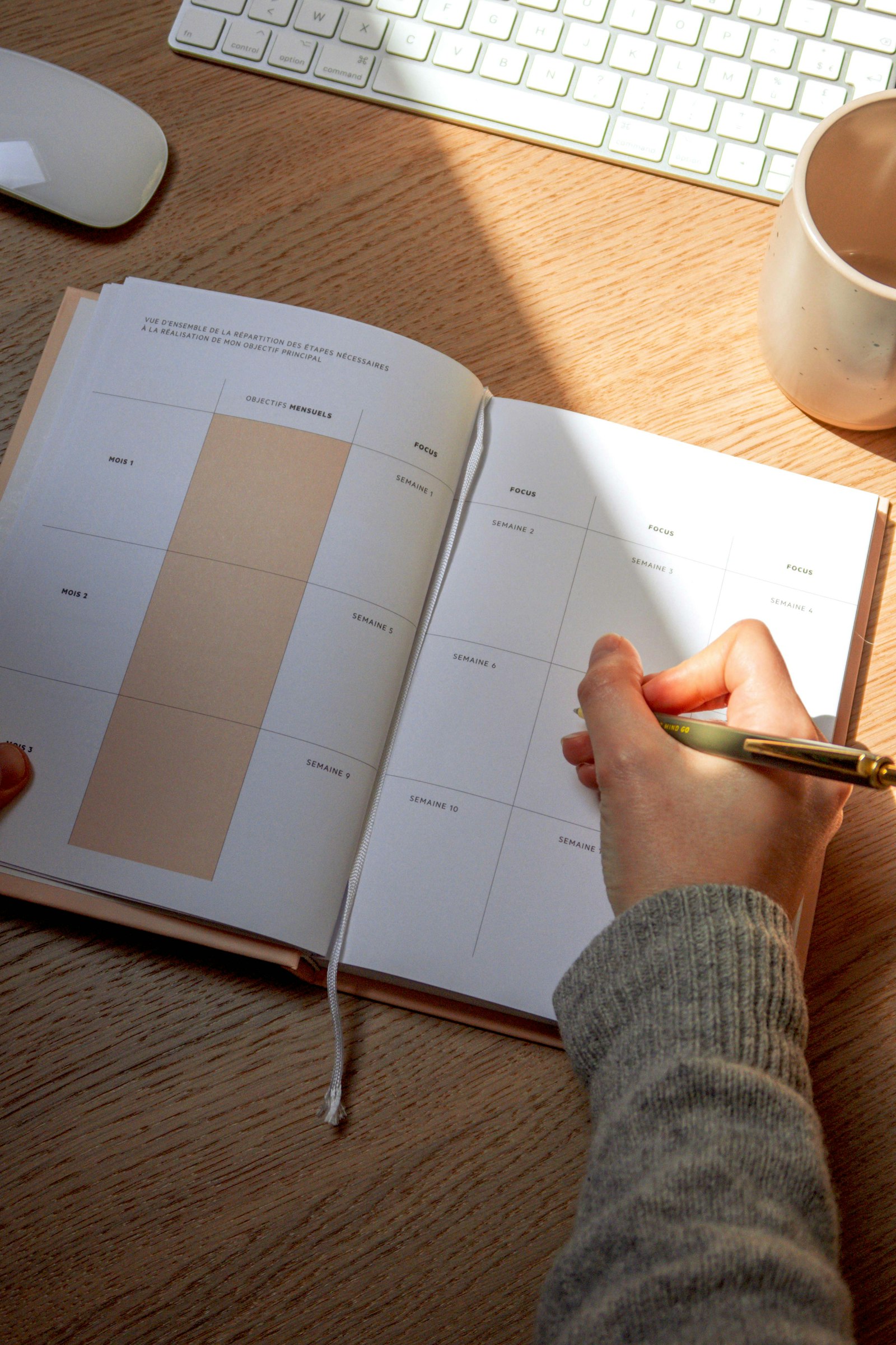 A hand writing in an open weekly planner on a wooden desk in warm daylight