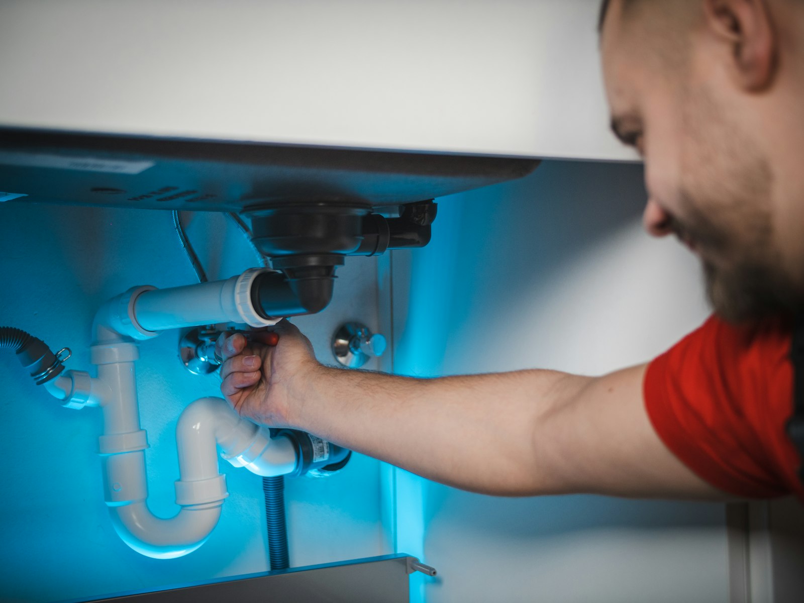 A plumber in a red shirt working on the pipes inside a wall beneath a sink