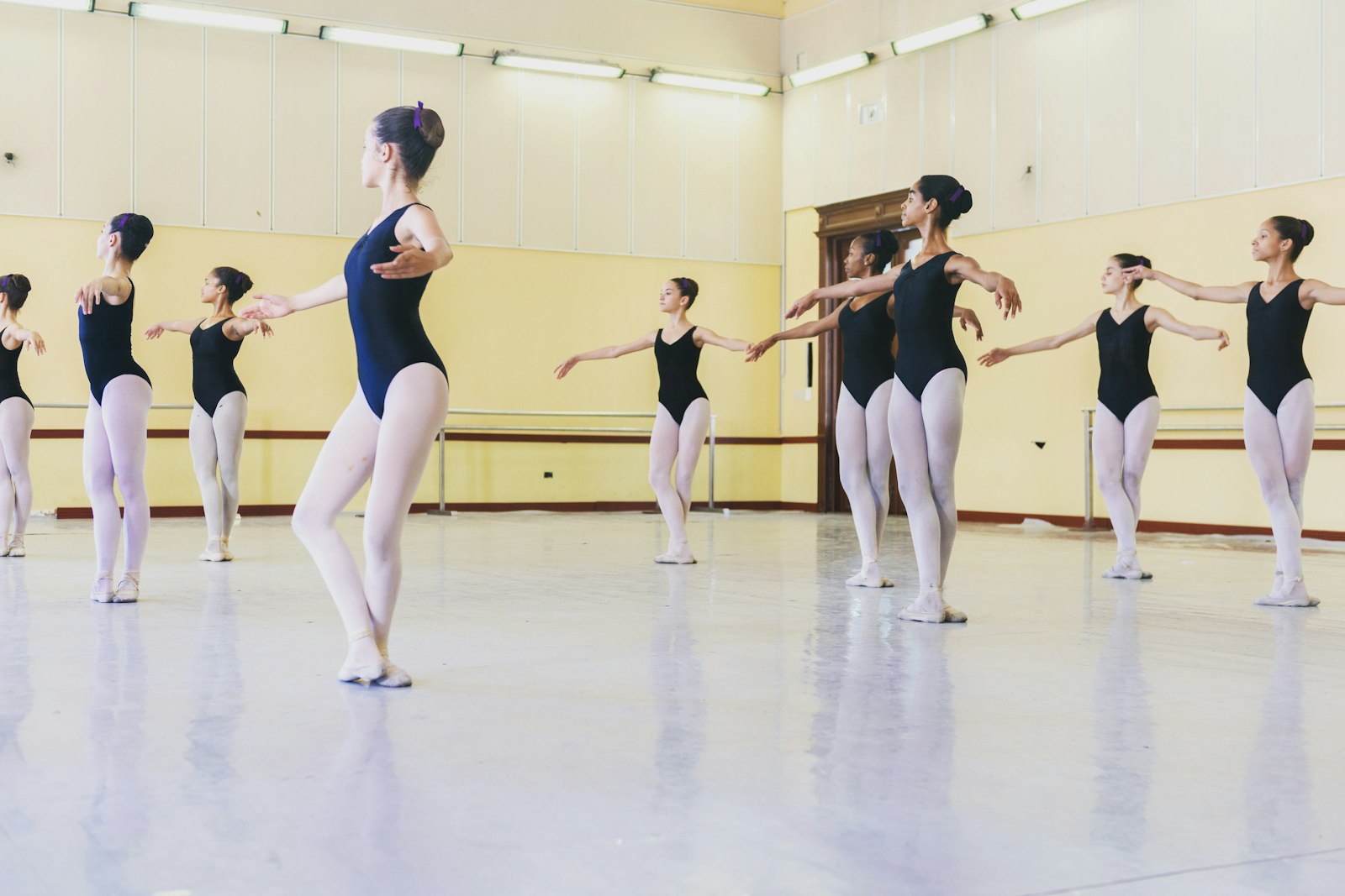Ballet class of students in black leotards holding an arm position on a white studio floor