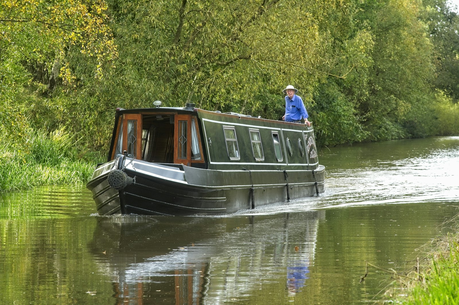 A green narrowboat cruising a tree-lined canal