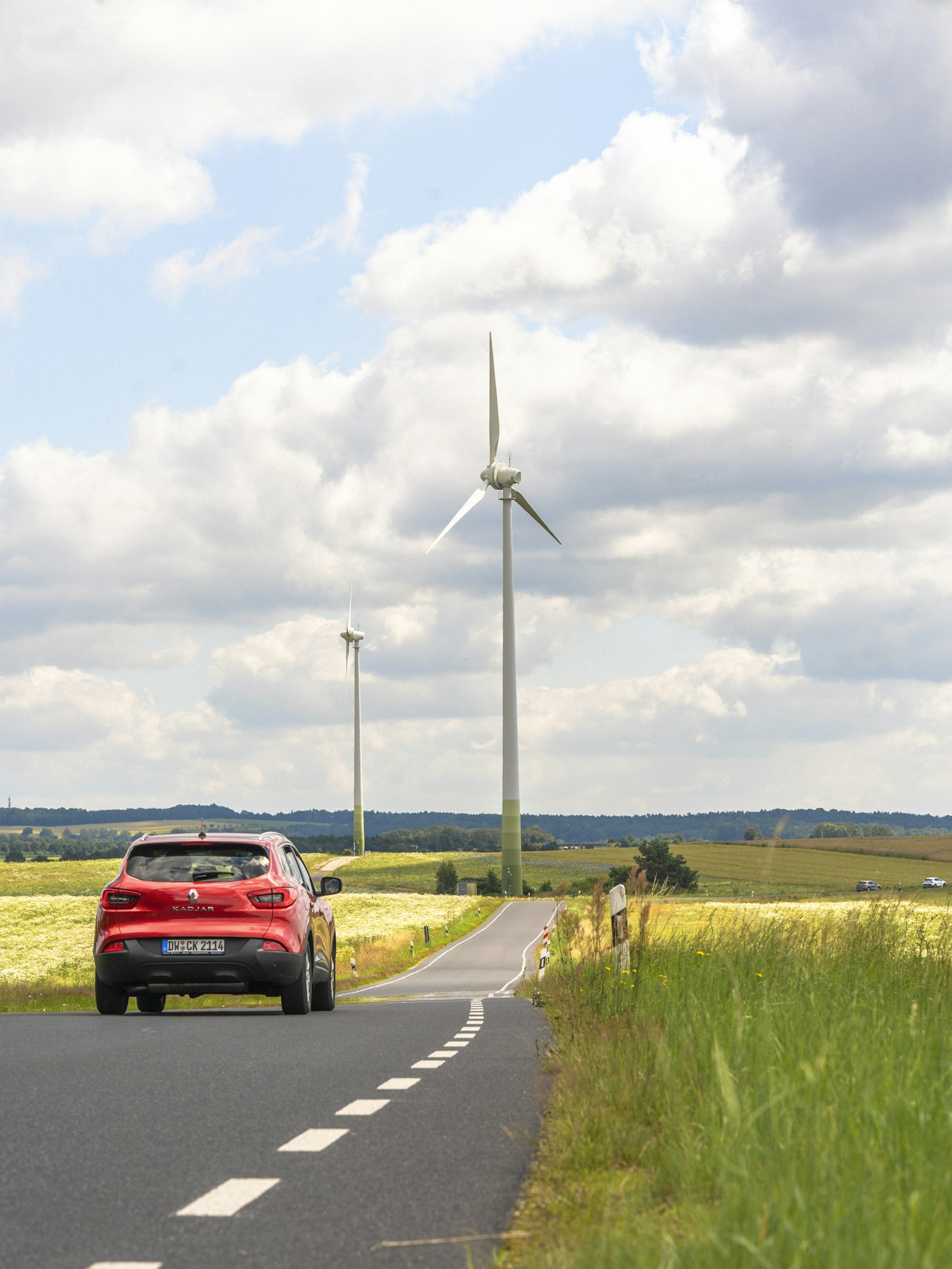 A red car on a narrow country road with a wind turbine beyond