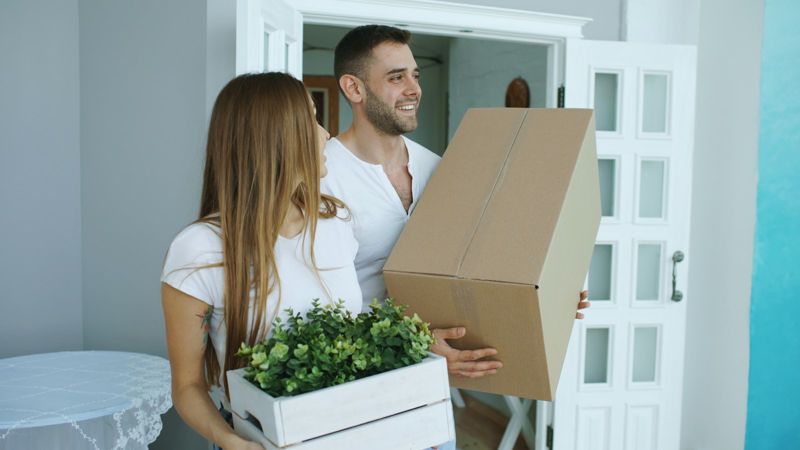A couple carrying a moving box and a plant through a doorway