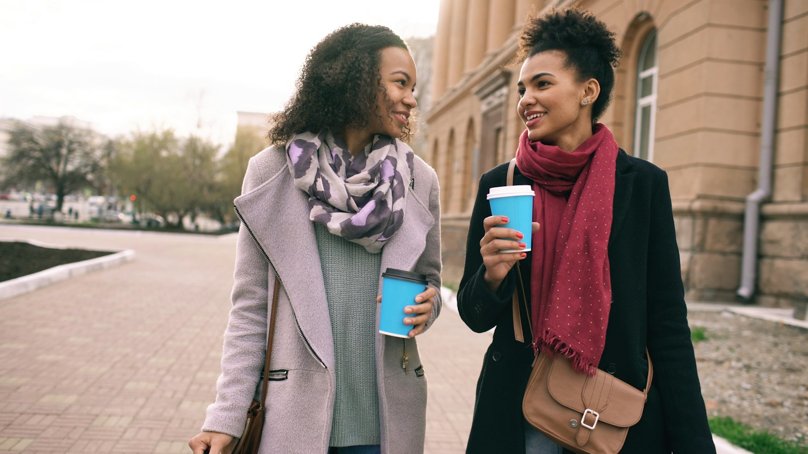 Two women walking side by side along a city pavement with takeaway coffees, mid-conversation