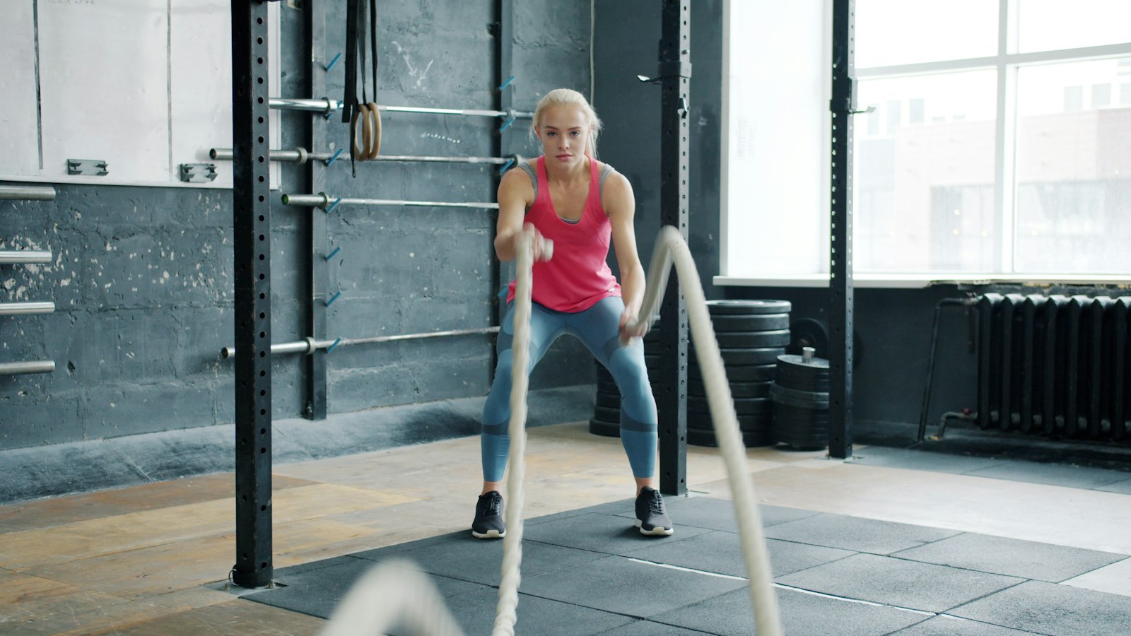 A woman working battle ropes in a bright gym with a rig behind her