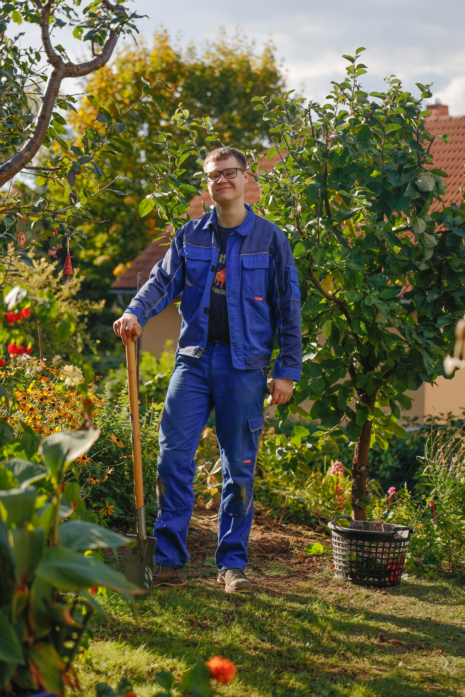 A gardener in blue overalls leaning on a spade under a fruit tree