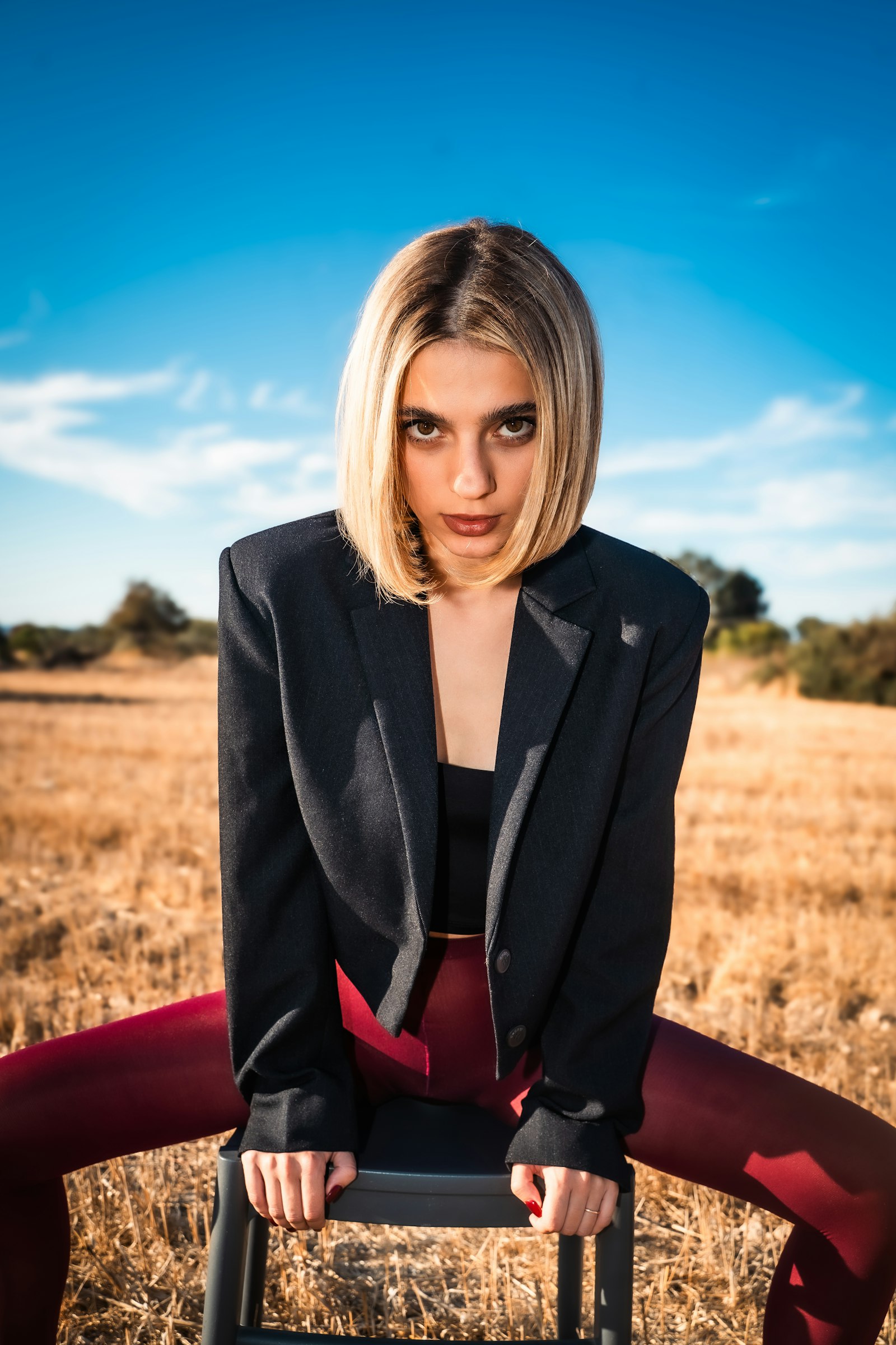 A woman in a black blazer leaning forward on a chair in a dry field under hard sun