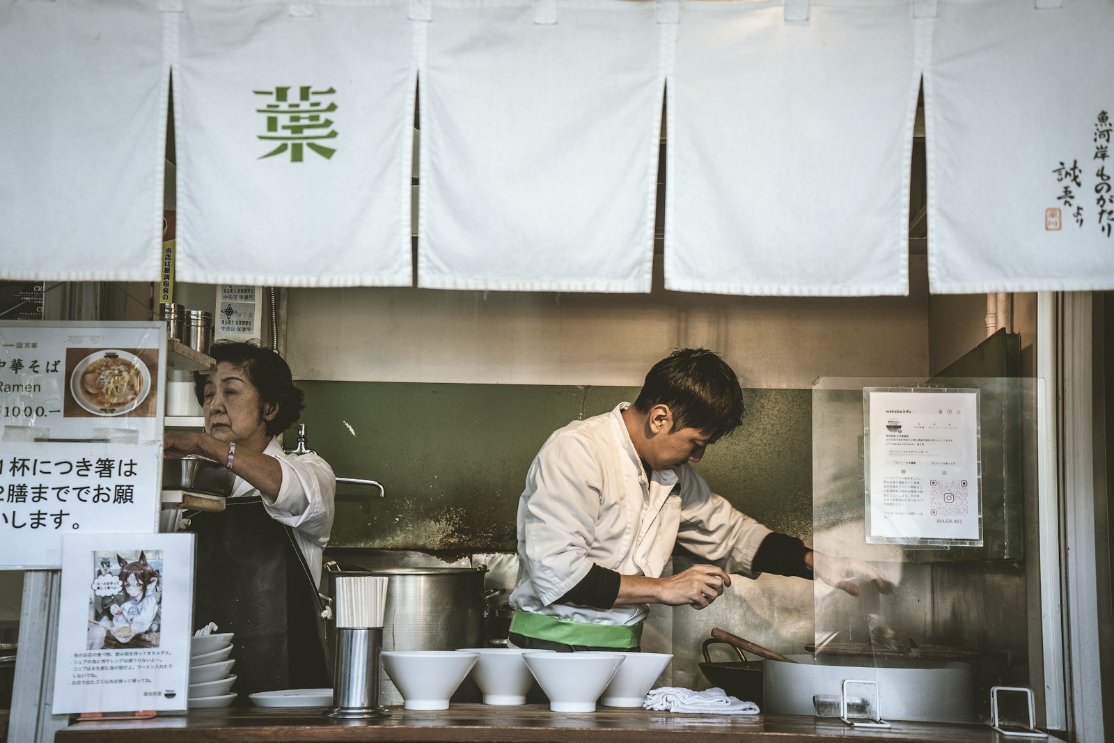 Two cooks at an open noodle kitchen behind white noren curtains